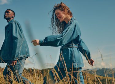 Na imagem temos um homem e uma mulher usando looks todo jeans, caminhando por um campo ao ar livre.