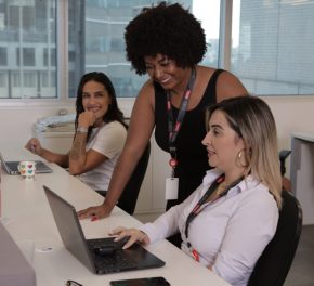 Na imagem vemos 3 mulheres no escritório interagindo em frente ao computador