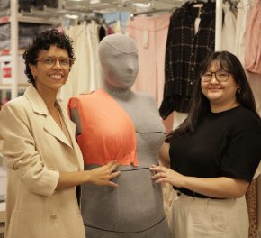 Na imagem vemos duas mulheres sorrindo ao lado de um manequim para modelagem de roupas