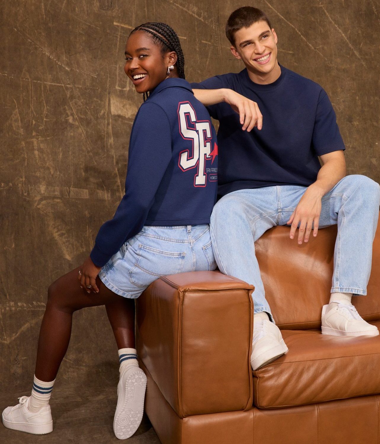 Dois jovens sentados em sofá. Mulher negra com moletom azul e shorts jeans. Homem branco com camiseta azul e calça jeans. Ambos sorrindo.